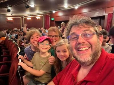 A smiling group of five people, including a bearded man in a red shirt and children, sit together in a theater. The audience is visible in the background.