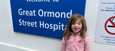 Girl in stripped pink top and multi coloured skirt with a black and white panda toy next to a a blue sign reading 'Welcome to Great ormond street hospital'
