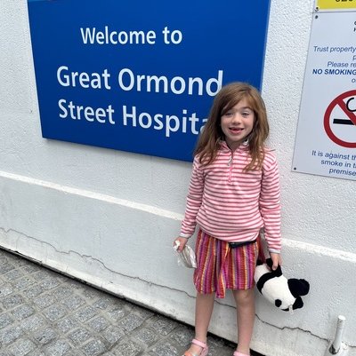 Girl in stripped pink top and multi coloured skirt with a black and white panda toy next to a a blue sign reading 'Welcome to Great ormond street hospital'