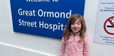 Girl in stripped pink top and multi coloured skirt with a black and white panda toy next to a a blue sign reading 'Welcome to Great ormond street hospital'