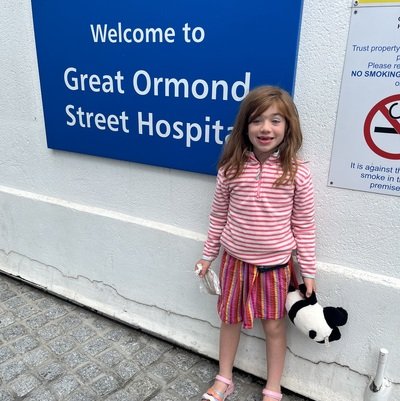 Girl in stripped pink top and multi coloured skirt with a black and white panda toy next to a a blue sign reading 'Welcome to Great ormond street hospital'