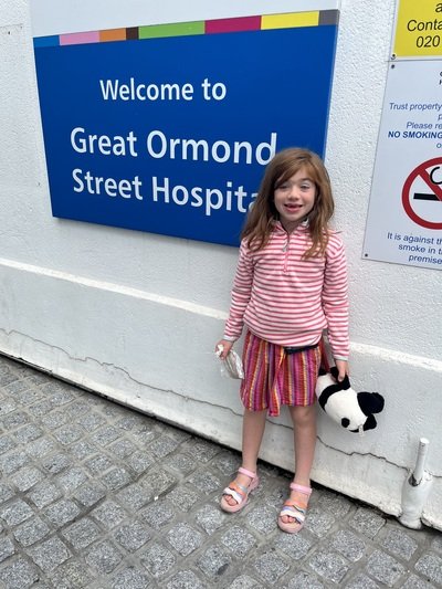 Girl in stripped pink top and multi coloured skirt with a black and white panda toy next to a a blue sign reading 'Welcome to Great ormond street hospital'