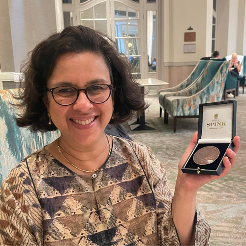 A woman in glasses smiles broadly at the camera while holding up a box with a large silver medal inside.