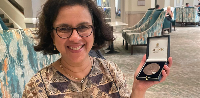 A woman in glasses smiles broadly at the camera while holding up a box with a large silver medal inside.