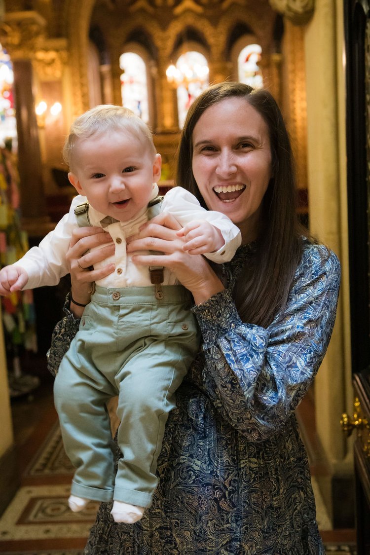 A woman joyfully holds a smiling baby in suspenders inside an ornately decorated building with warm lighting and stained-glass windows in the background.