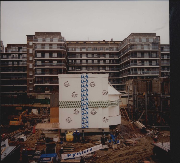 Construction site with a large building in the background, featuring art deco design. Workers and machinery are in the foreground amid scaffolding.