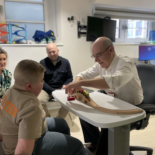 A man with a white shirt and sleeves rolled up plays with trains at a table with a young boy. Two adults are smiling in the background and it looks like they are his parents