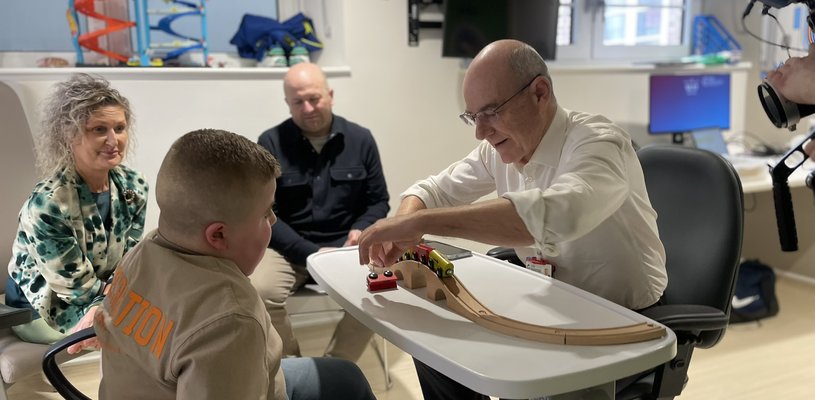 A man with a white shirt and sleeves rolled up plays with trains at a table with a young boy. Two adults are smiling in the background and it looks like they are his parents
