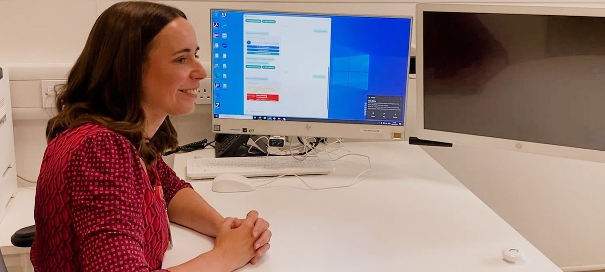 Female clinician sat at desk in clinic room smiling and looking towards patient. Computer screen in background.