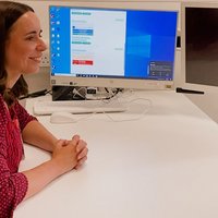 Female clinician sat at desk in clinic room smiling and looking towards patient. Computer screen in background.