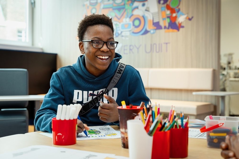 A young man in a hoodie is sat at a table smiling broadly. The table is covered in colouring sheets, pens and pencils