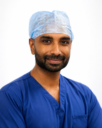 A man smiling at the camera. He is wearing blue medical scrubs.