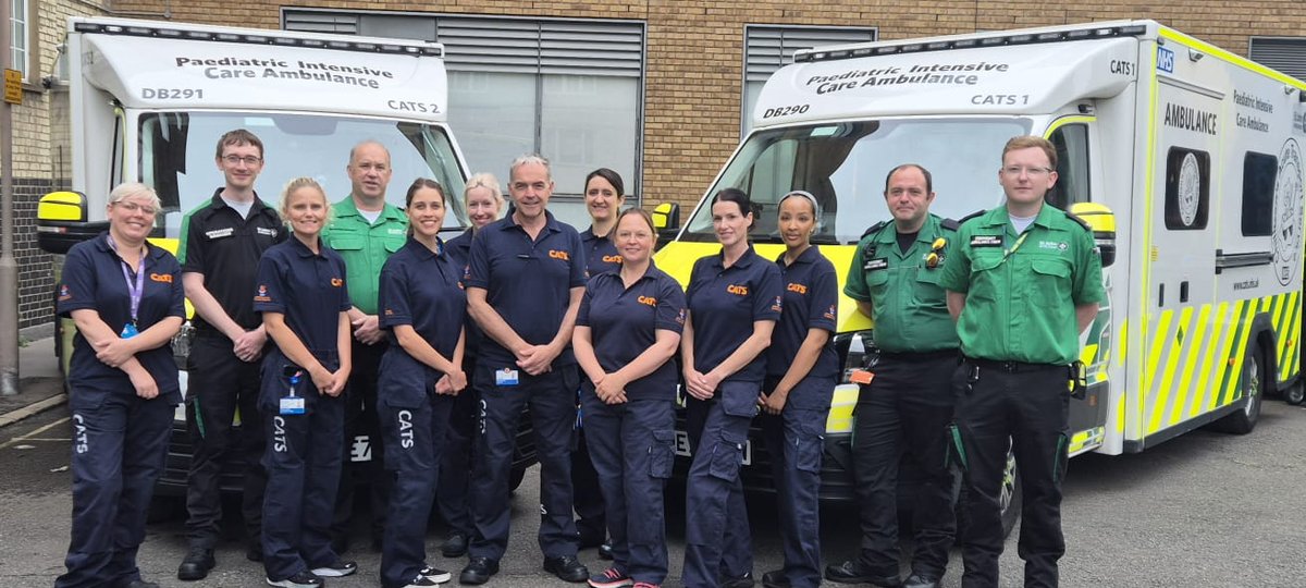 A group of medical professionals in paramedic uniforms stand smiling in front of two pediatric intensive care ambulances. They convey teamwork and readiness.