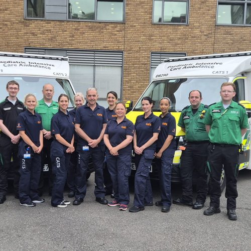 A group of medical professionals in paramedic uniforms stand smiling in front of two pediatric intensive care ambulances. They convey teamwork and readiness.