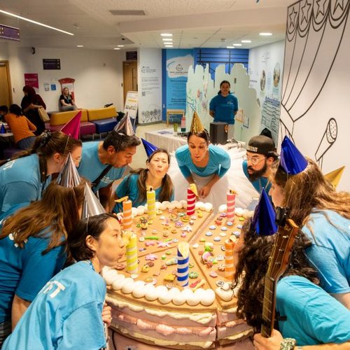 A group of people are stood around a giant cake and they are blowing out the candles