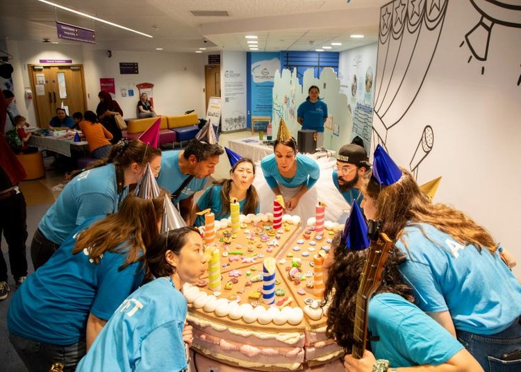 A group of people are stood around a giant cake and they are blowing out the candles