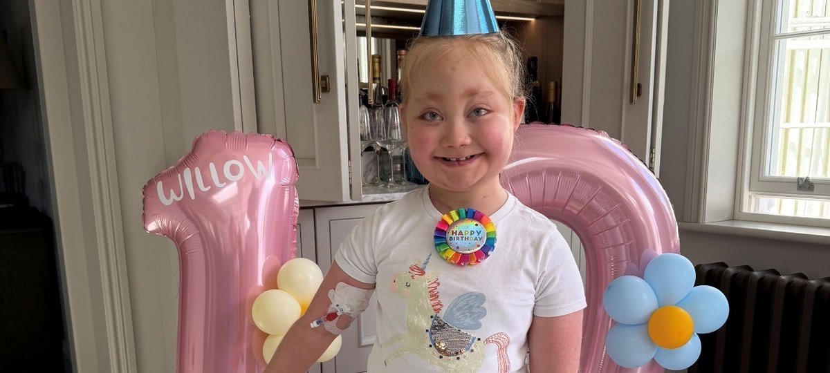 A young girl in a blue party hat smiles broadly in front of balloons that are in the shape of the number 10