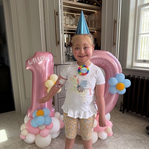 A young girl in a blue party hat smiles broadly in front of balloons that are in the shape of the number 10