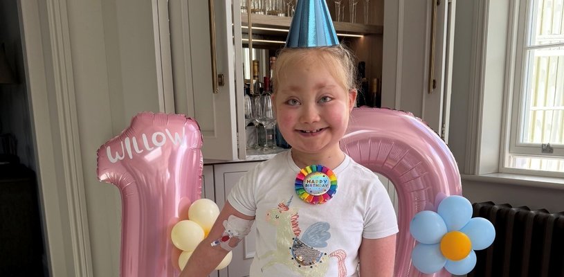 A young girl in a blue party hat smiles broadly in front of balloons that are in the shape of the number 10