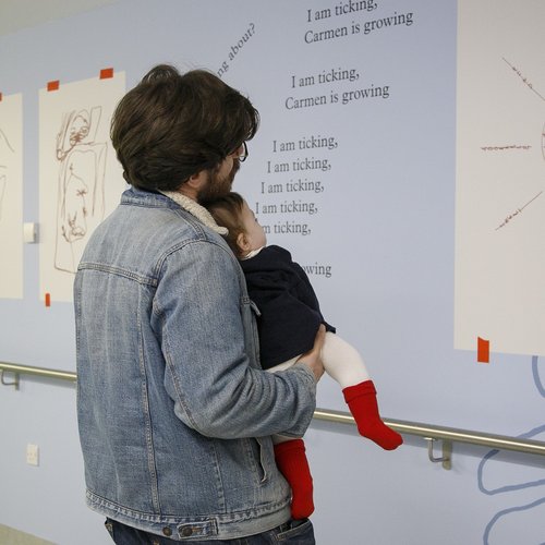 A man holding a baby looking at a sun artwork on the wall on a hospital corridor
