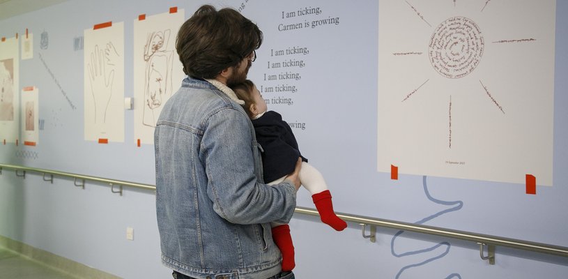 A man holding a baby looking at a sun artwork on the wall on a hospital corridor