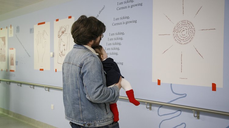 A man holding a baby looking at a sun artwork on the wall on a hospital corridor