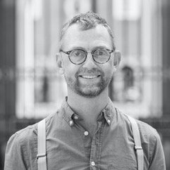 A black and white photo of a man smiling - he has a beard and is wearing glasses