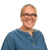 A woman with blonde hair, black glasses, wearing a blue top is smiling.