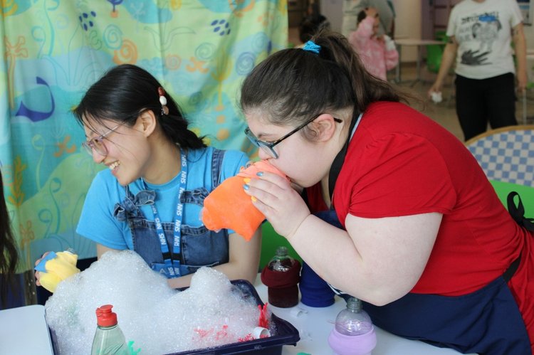 Two women sat at a table with a washing up bowl on a table with water and bubbles in it