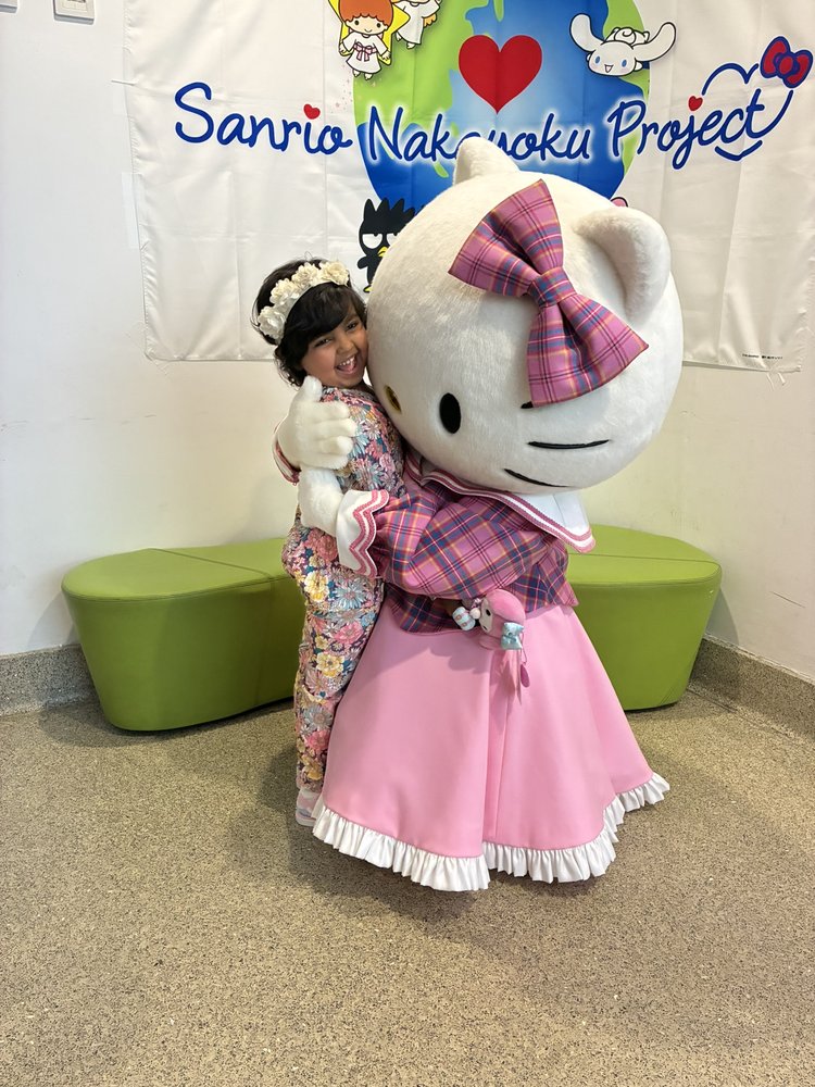 Girl in floral outfit with flower crown and black hair hugging hello kitty character with a green and white background