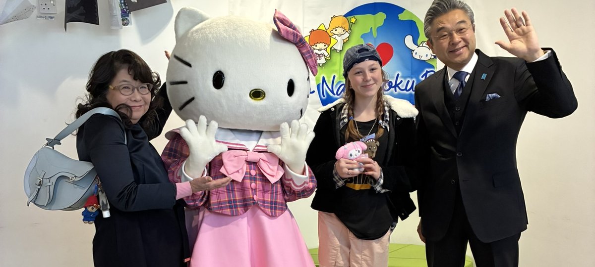 Two women and a man happily pose with a Hello Kitty mascot in a pink dress. The setting is cheerful, with a colorful backdrop.
