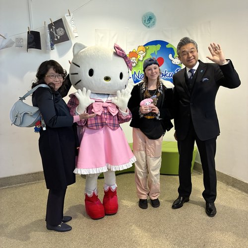 Two women and a man happily pose with a Hello Kitty mascot in a pink dress. The setting is cheerful, with a colorful backdrop.