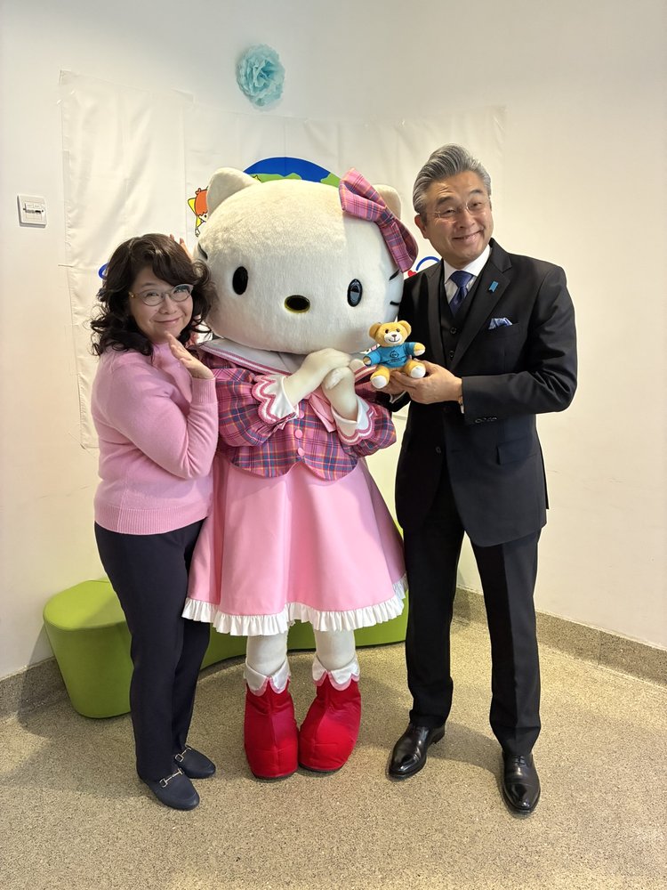 A woman in pink and a man in a suit with a teddy bear pose alongside a large character in a pink dress, conveying a joyful and whimsical atmosphere.