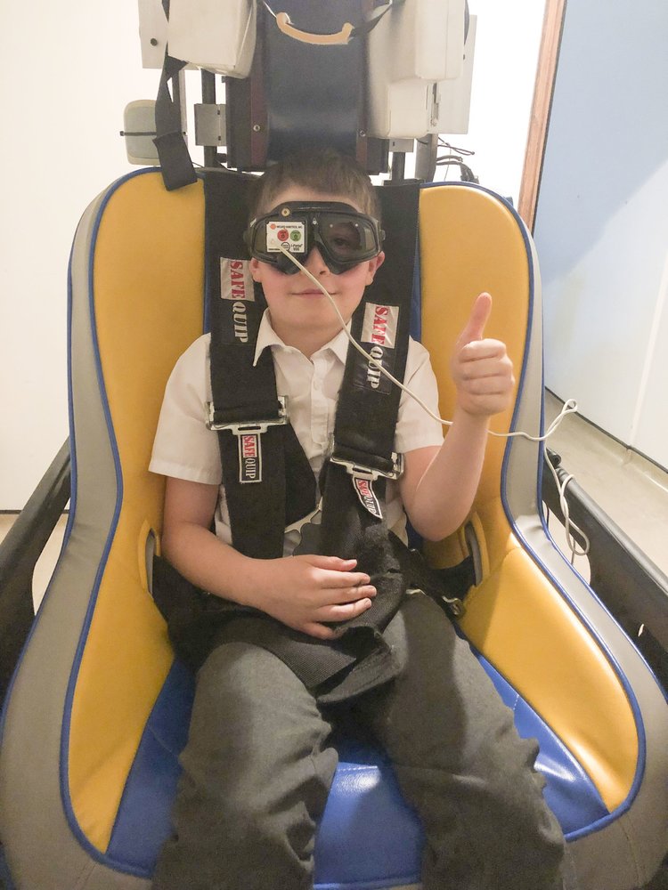 A child is sat in a chair with a belt on, they are wearing googles and have their thumb up.