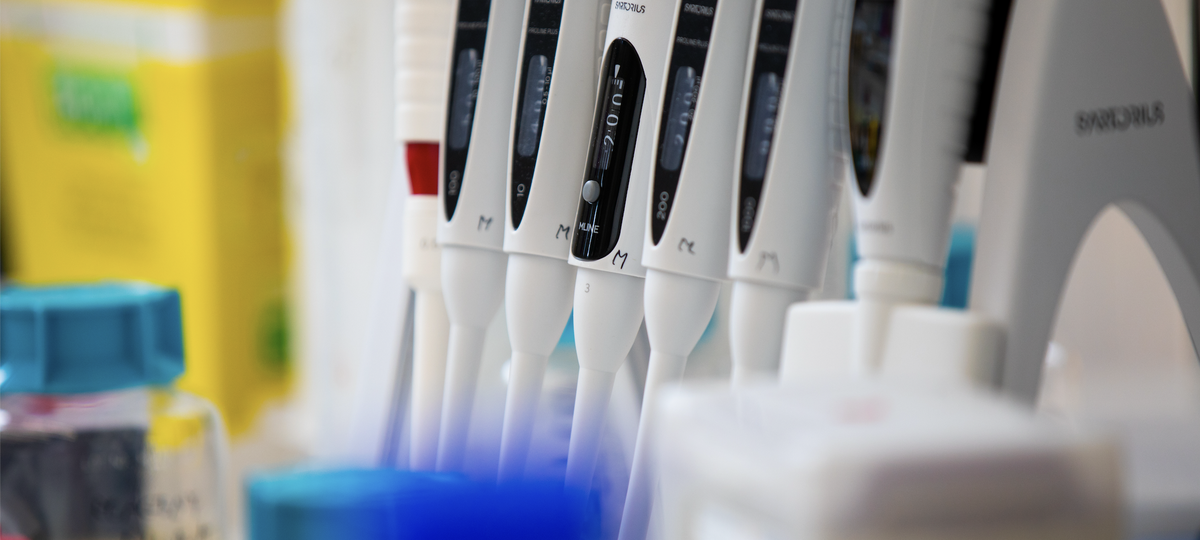 a close-up view of several adjustable laboratory pipettes in a holder, with lab containers in the background.