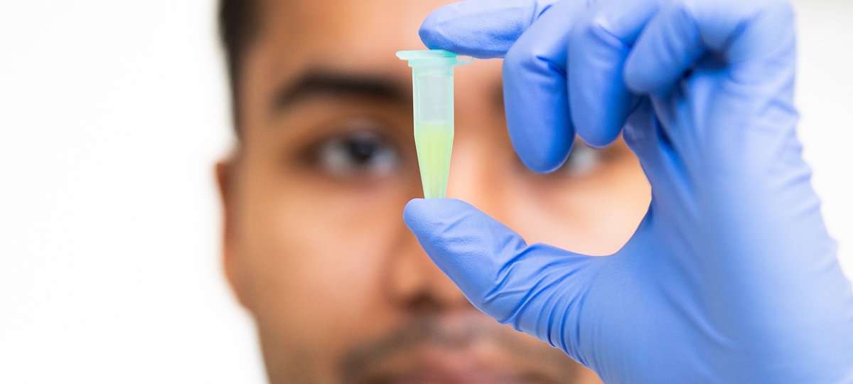 A close up image of a vial of liquid with a blurry outline of the man holding it behind