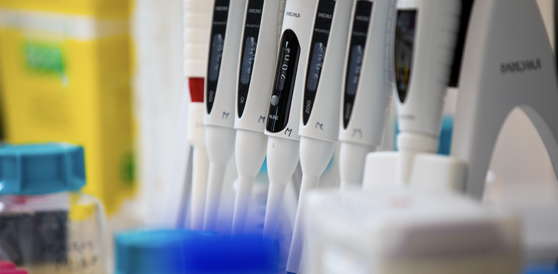 a close-up view of several adjustable laboratory pipettes in a holder, with lab containers in the background.