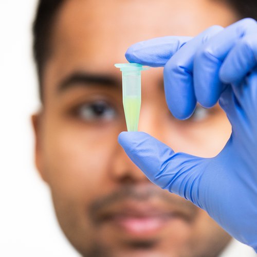 A close up image of a vial of liquid with a blurry outline of the man holding it behind