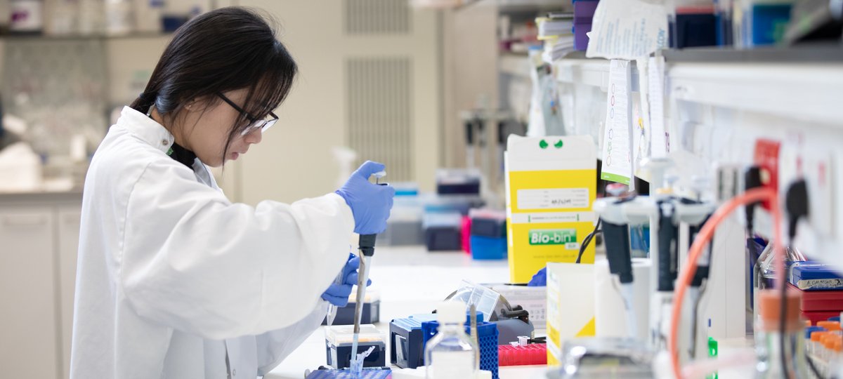 Researcher wears white lab coat and uses pipette in lab.