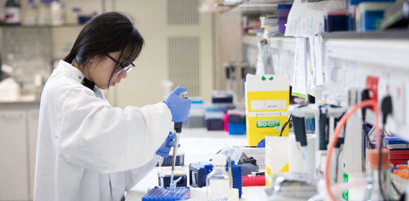 Researcher wears white lab coat and uses pipette in lab.