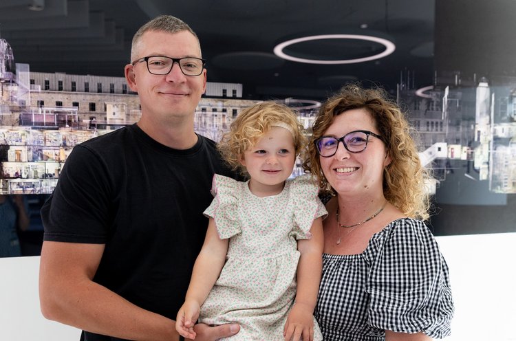 Dad Lukasz and mum Sandra holding daughter Phoebe in the middle, standing in front of a black photograph.