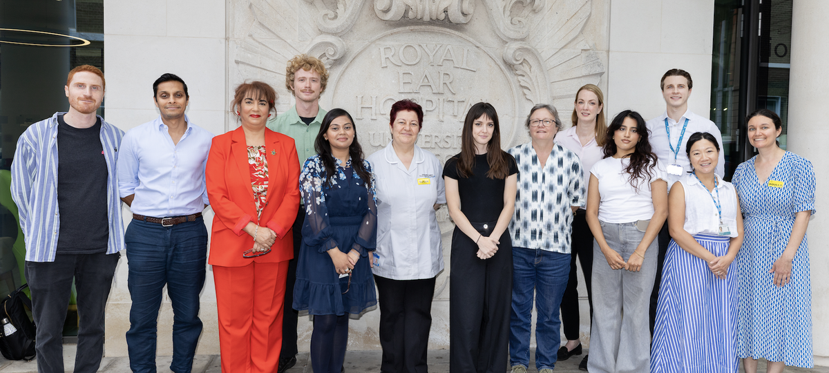 13 staff members, some wearing uniform and some wearing their own clothes, stood in front of the sign for the Royal Ear Hospital the