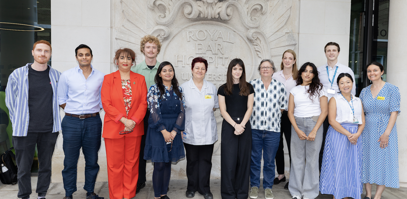 13 staff members, some wearing uniform and some wearing their own clothes, stood in front of the sign for the Royal Ear Hospital the