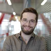 A bearded white man with brown hair smiles at the camera. He is wearing silver framed circular glasses