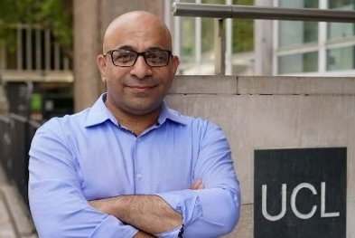 A man with glasses leans against a wall next the a sign saying UCL