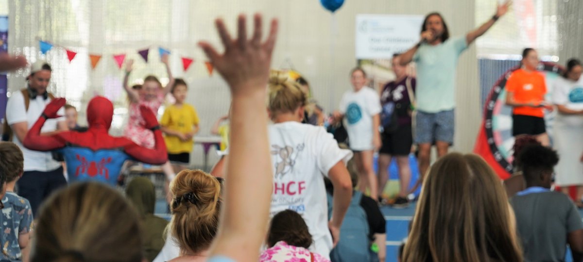A out of focus image looking at Joe Wicks doing a warm up. Joe is stood at the front, with an arm in the air, and there are children either side of him. You can see the backs of children's heads who are taking part in the warm up.