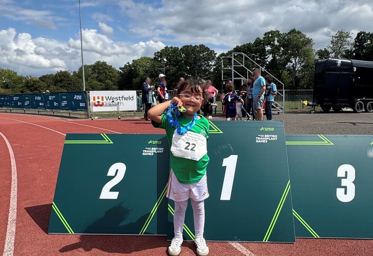 A young patient is standing proudly on a race track, in front of a sports podium, holding up a medal. They have black hair and pink glasses, and are wearing a green top.