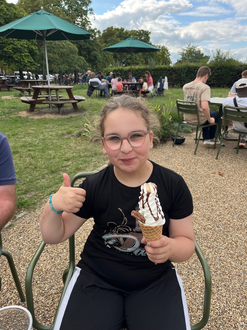A young girl holds up an ice cream and gives a thumbs up to the camera. She is smiling widely.