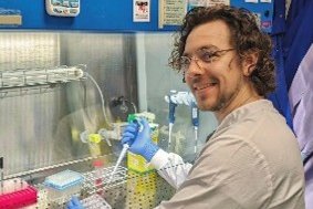A man in glasses sits at a lab bench and wears a lab coat and blue gloves. he is smiling at the camera using lab equipment
