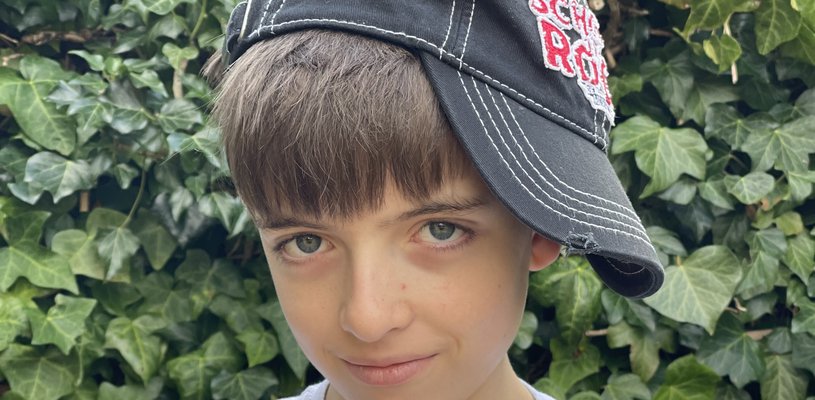 12 year old Oliver wears a School of Rock baseball cap, tilted to the side, with his arms crossed and his hands in a V sign.